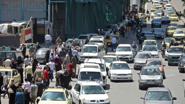 In this file photo taken on May 10, 2020, pedestrians walk along a line of cars in a busy street in the Syrian capital Damascus