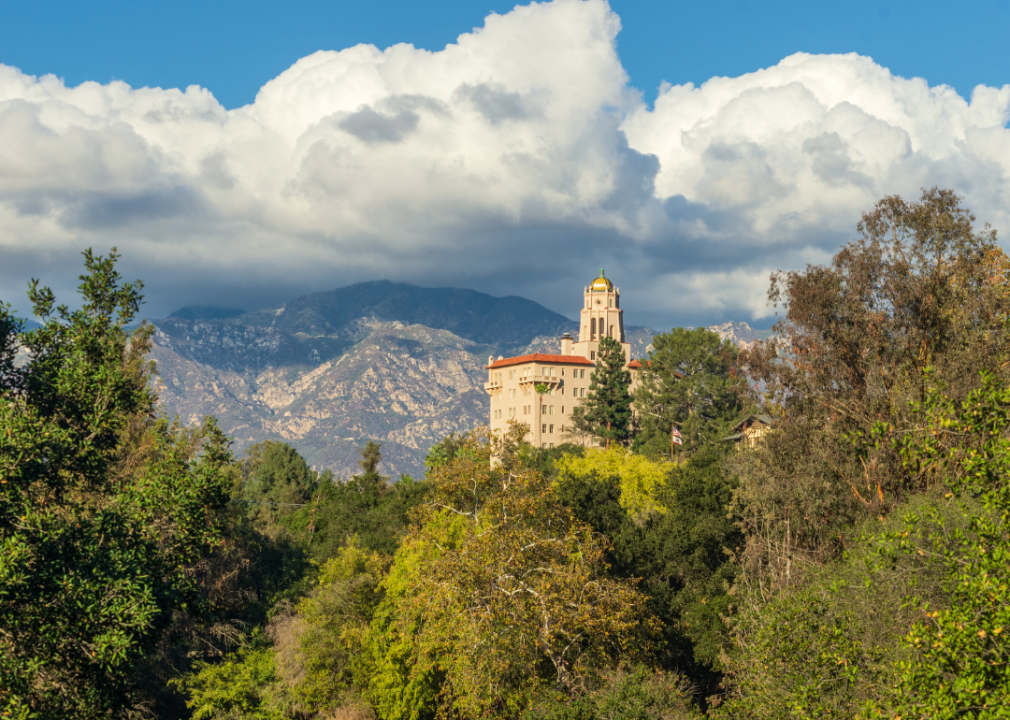 A large, white building with a clock tower surrounded by trees and mountains. 